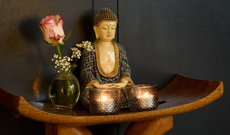 een boeddhabeeldje zit op een houten krukje met ernaast een roos in een vaasje en ervoor twee kaarsjes
